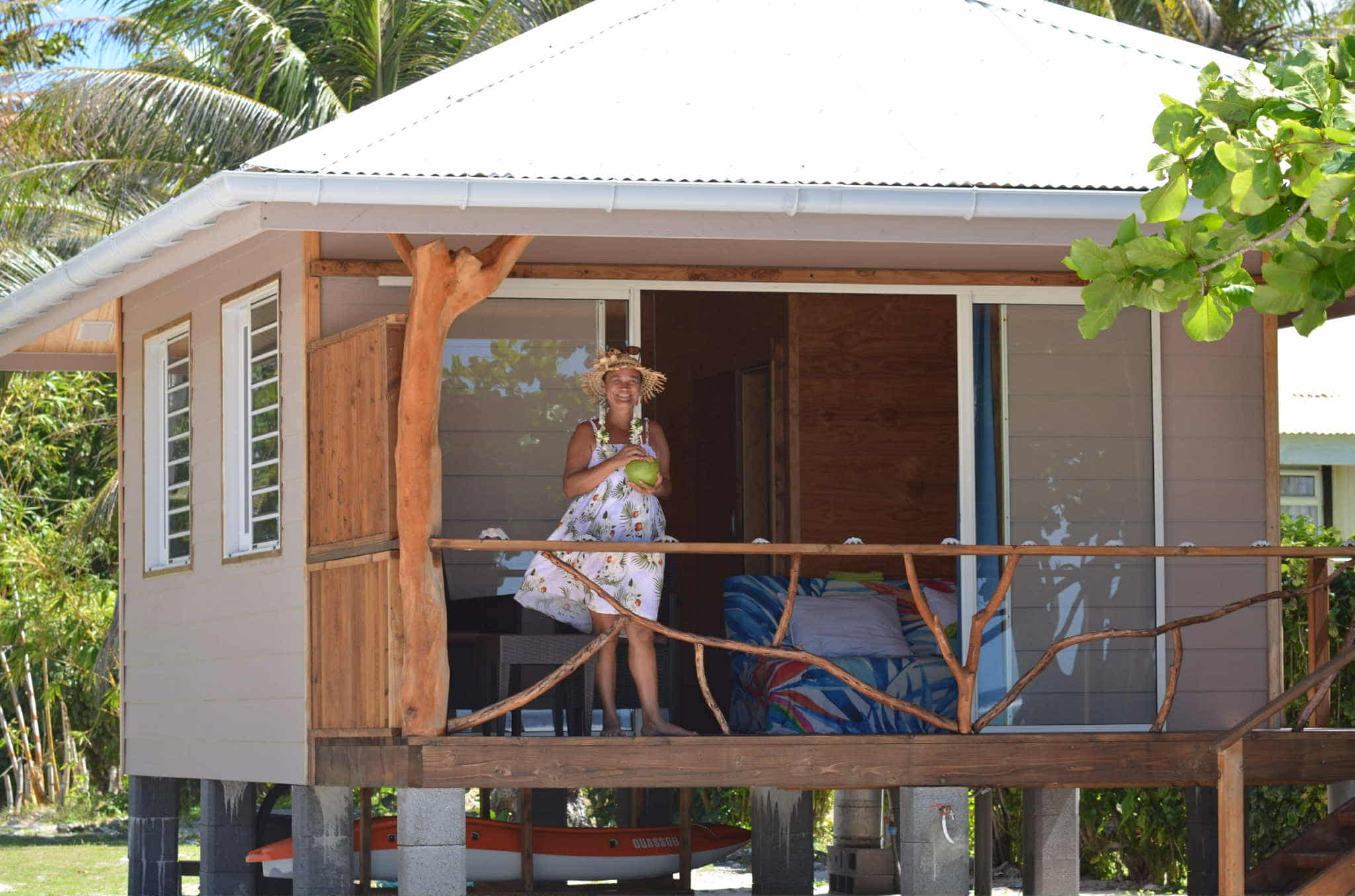 Hôtesse en robe à fleurs et couronne, une noix de coco à la main, sur la terrasse en bois d'un bungalow de Tiahani Lodge